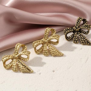 May include: Three bow-shaped brooches in gold and bronze tones. The brooches have a textured surface with small, raised dots. One brooch has the text "customaffordable" on it. The brooches are arranged on a white surface with a pink satin fabric.