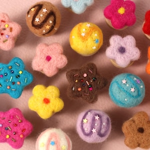May include: A collection of colorful, felted wool ornaments in various shapes, including stars, flowers, and round shapes. The ornaments are decorated with beads and sprinkles, and are in shades of pink, blue, yellow, brown, and orange.
