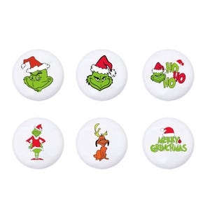 Op de afbeelding: Zes witte ronde keramische ornamenten met verschillende kerstthema's met de Grinch. Eén ornament heeft de Grinch met een kerstmanmuts en de tekst "Ho Ho Ho". Een ander ornament heeft de Grinch met een kerstmanmuts en de tekst "Merry Grinchmas". Een ander ornament heeft de Grinch met een kerstmanmuts en een chagrijnige blik. Een ander ornament heeft de Grinch met een kerstmanmuts en een cadeau in zijn hand. Een ander ornament heeft de Grinch met een kerstmanmuts en een hond. Het laatste ornament heeft de Grinch met een kerstmanmuts en een rendier.