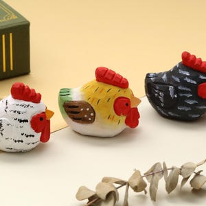Puede incluir: Tres figuras de gallina de madera pintadas a mano en blanco, amarillo y negro. Cada una tiene una cresta y una barbilla rojas, con un pico amarillo. Las gallinas están dispuestas sobre una superficie amarillo claro, lo que sugiere un tema decorativo o de colección.