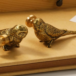 May include: Two brass bird-shaped cabinet knobs. The knobs are detailed with feathers and have a threaded post for attachment. The birds are positioned on a light brown surface, suggesting a decorative or functional item for furniture.