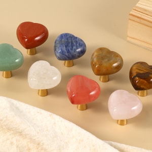May include: A selection of heart-shaped cabinet knobs in diverse colours and textures. The knobs are crafted from polished gemstones, such as red jasper, green aventurine, and rose quartz. Each knob features a gold-coloured base.