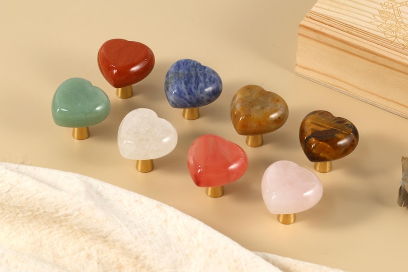 May include: A selection of heart-shaped cabinet knobs in diverse colours and textures. The knobs are crafted from polished gemstones, such as red jasper, green aventurine, and rose quartz. Each knob features a gold-coloured base.