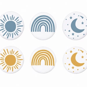Könnte beinhalten: Sechs weiße runde Knöpfe mit Himmelsdesigns. Die Designs umfassen eine Sonne, einen Regenbogen und einen Mond mit Sternen. Sonne und Regenbogen sind blau und gelb. Mond und Sterne sind blau und gelb.