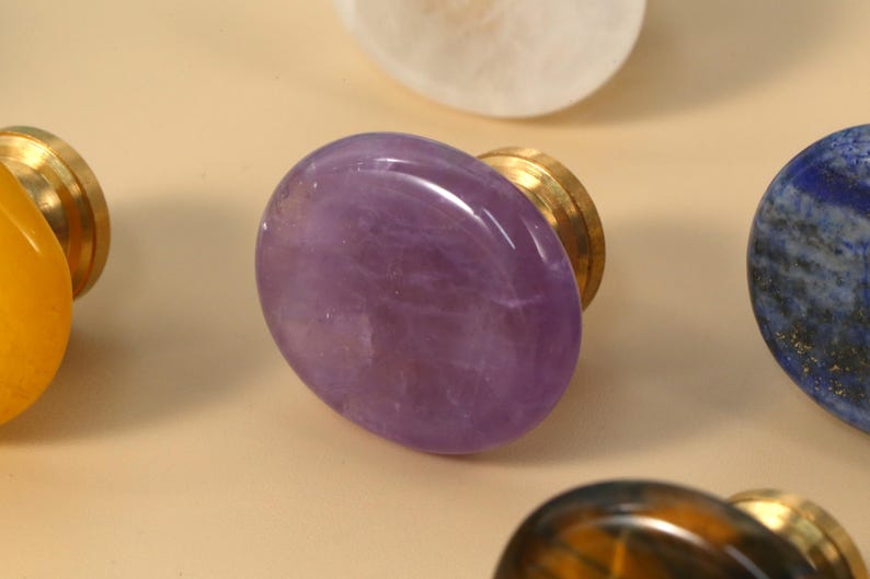 May include: A selection of gemstone cabinet knobs. The knobs have oval-shaped polished stones in various colours, including purple, yellow, white, blue, and brown, set on brass bases. The amethyst knob is in focus.