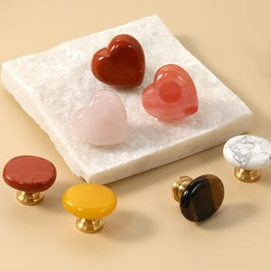 May include: A selection of decorative cabinet knobs in various colours and shapes. The knobs include heart-shaped stones and round knobs in red, pink, yellow, and marble patterns. Each knob has a gold-coloured base.