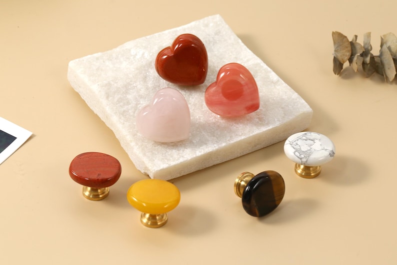 May include: A selection of decorative cabinet knobs in various colours and shapes. The knobs include heart-shaped stones and round knobs in red, pink, yellow, and marble patterns. Each knob has a gold-coloured base.