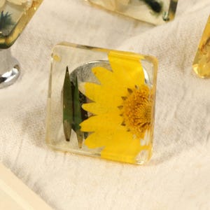 Op de afbeelding: Transparante vierkante kastknoppen met een gele zonnebloem ingekapseld in hars. De knoppen hebben een zilverkleurige metalen basis. Andere knoppen met gele en groene bloemmotieven zijn ook zichtbaar.