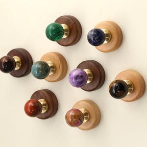 Pode incluir: Uma coleção de puxadores decorativos com esferas de pedra polida coloridas. Os puxadores são montados em bases redondas de madeira com ferragens de latão. Os tons de madeira variam de claros a escuros, e as cores das pedras incluem verde, azul, roxo e vermelho.