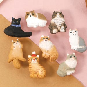 Op de afbeelding: Een verzameling handgeschilderde kattenbeeldjes in verschillende poses en kleuren. De katten zijn in tinten zwart, bruin, wit en grijs, sommige met accessoires. De beeldjes zijn gerangschikt op een roze en bruin oppervlak.