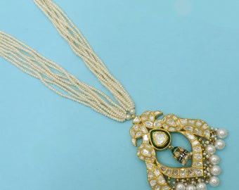 Puede incluir: Un collar de oro con un gran colgante adornado con una piedra preciosa verde y perlas blancas. El colgante está rodeado de perlas blancas más pequeñas y tiene un diseño delicado.