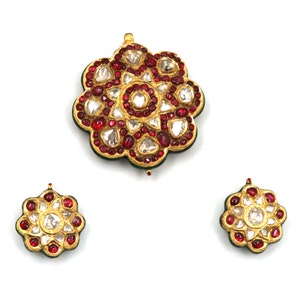 May include: A set of gold jewelry featuring a large pendant and two earrings. The pendant and earrings are shaped like flowers with a green background and are adorned with red and white gemstones.