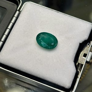 Puede incluir: Una gema de esmeralda verde vibrante, de forma ovalada, colocada en una caja de joyería de terciopelo blanco.