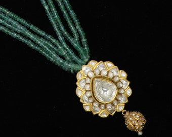 Puede incluir: Un collar de oro con un gran colgante de diamante en forma de pera rodeado de diamantes más pequeños. El collar está hecho de cinco hileras de cuentas de color verde esmeralda.