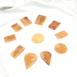 Puede incluir: Una colección de piedras preciosas pulidas en varias formas y tonos de naranja y amarillo. El surtido incluye piedras rectangulares, ovaladas, en forma de lágrima y circulares, dispuestas sobre una superficie blanca.