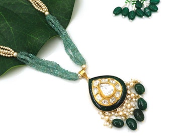 Puede incluir: Un collar con un colgante en forma de lágrima de color verde y dorado, adornado con una piedra grande transparente rodeada de piedras más pequeñas. El colgante está unido a un collar multicadena de cuentas azul claro.