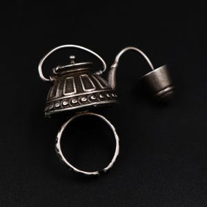 May include: A silver ring featuring a teapot design. The teapot has a spout and a lid, with a small cup attached to the spout.