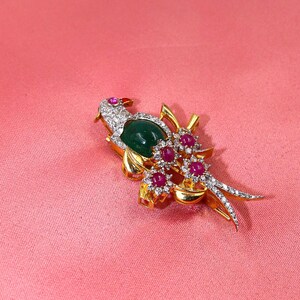 May include: A gold-toned bird brooch with a large oval green stone body, ruby accents, and diamond-like detailing. The bird's head features a small ruby eye. The brooch is set against a pink background.