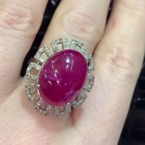 Könnte beinhalten: Ein Ring mit einem großen ovalen Rubin, der von einem Heiligenschein aus kleineren Diamanten umgeben ist. Der Ring ist in Silber gefasst.