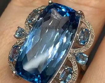 May include: A large, rectangular, light blue gemstone ring with a silver setting. The ring features smaller blue stones and clear accent stones around the main stone. The ring is on a finger.