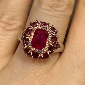 Könnte beinhalten: Ein silberner Ring mit einem großen, rechteckigen, roten Rubin-Edelstein, umgeben von kleineren roten Edelsteinen und einigen kleinen, klaren Edelsteinen.