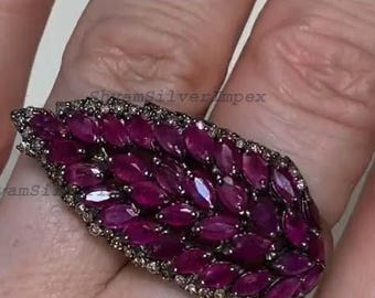 May include: A statement ring with a leaf design, set with numerous oval-cut, deep red gemstones. The ring has a dark metal band, with smaller, clear stones outlining the leaf shape. The ring is worn on a finger.