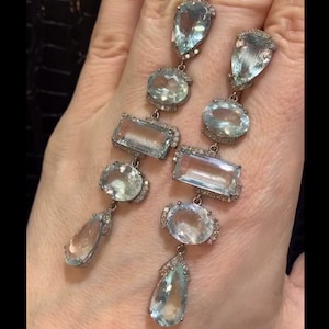 May include: A pair of long, silver dangle earrings featuring multiple aquamarine gemstones. The earrings have a variety of shapes, including pear, oval, and rectangular. The gemstones are set in silver and have a clear, light blue colour.