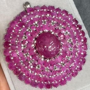 Könnte beinhalten: Ein großer, kreisförmiger Anhänger mit einem zentralen ovalen Rubin, umgeben von blütenförmigen Rubinen. Der Anhänger hat eine silberfarbene Öse. Der Text "shyamsilverimpex" ist sichtbar. Ein luxuriöses Schmuckstück.