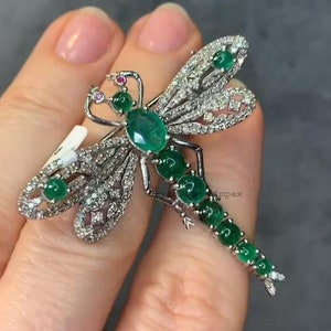 Puede incluir: Un broche de libélula de plata con piedras de esmeralda verde y diamantes transparentes. Las alas de la libélula están extendidas y el cuerpo está adornado con piedras de esmeralda.
