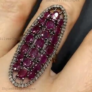 Könnte beinhalten: Ein ovaler Ring mit einem Cluster aus oval geschliffenen, tiefroten Edelsteinen. Die Edelsteine sind von einem Rand aus kleinen, klaren Steinen umgeben. Der Ring ist auf einem silbernen Band gefasst.
