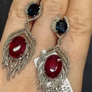 Peut inclure: Une paire de boucles d'oreilles pendantes avec un motif de pierres précieuses rouges et bleues. Les boucles d'oreilles présentent une grande pierre précieuse ovale rouge entourée de diamants plus petits, et une plus petite pierre précieuse ovale bleue en haut. Les boucles d'oreilles sont en argent et ont un design en forme de plume.