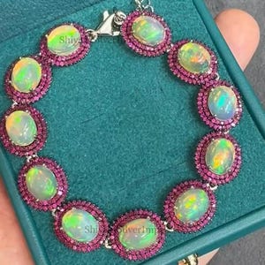 May include: An opal bracelet with oval-shaped opals surrounded by small, round, pink gemstones. The bracelet is set in a silver metal and is displayed in a teal jewelry box. The text "Shiy SilverImp" is visible.