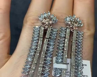 May include: A pair of silver-toned drop earrings. Each earring features a floral design at the top and a rectangular shape with rows of light blue and clear crystals. The earrings are held in a hand.