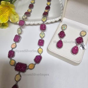 May include: A necklace and earring set featuring ruby and opal gemstones. The necklace has a series of ruby and opal stones set in a silver metal. The earrings mirror the necklace design. The gemstones are set in a silver metal with a diamond-like border.