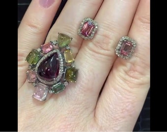 May include: A ring with a large, teardrop-shaped, dark purple gemstone surrounded by smaller, multi-colored gemstones. The ring is set in silver and has a delicate, intricate design. Two matching earrings with pink gemstones and diamond accents are also shown.