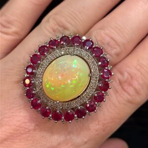 Op de afbeelding: Een ovale ring met een grote, iriserende opaal in het midden, omringd door kleine, ronde robijnen. De ring heeft een zilverkleurige band en een rand van kleine, heldere edelstenen. De opaal vertoont een kleurenspel.