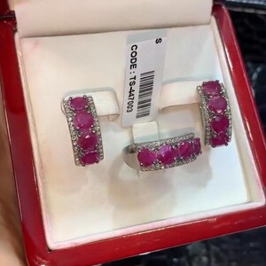Puede incluir: Un conjunto de joyas con un anillo y un par de pendientes, cada uno adornado con gemas rojas vibrantes y pequeñas piedras claras. El conjunto se presenta en una caja roja con interior blanco. La etiqueta dice "CODE: TS-447003".