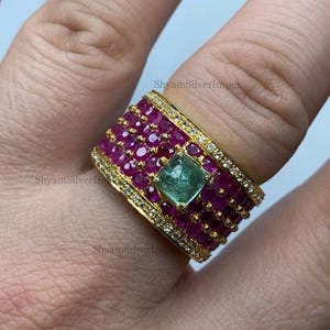 Peut inclure: Une bague dorée ornée d'un émeraude carrée centrale, entourée de petits rubis ronds rouge foncé. Les bords de la bague sont ornés de petits pierres précieuses transparentes. Un bijou remarquable.