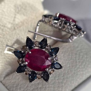 Könnte beinhalten: Silberne Ohrringe mit einem zentralen ovalen Rubin, umgeben von dunkelblauen Saphiren und kleinen Diamanten. Die Ohrringe haben ein florales Design und einen Klappverschluss. Der Markenname "ShyamSilverImpex" ist sichtbar.