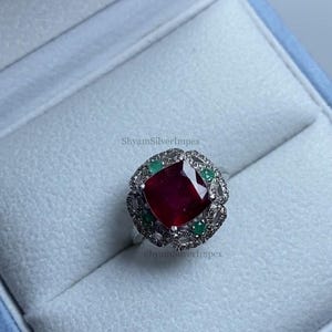 Könnte beinhalten: Ein silberner Ring mit einem großen, quadratisch geschliffenen, tiefroten Rubin im Zentrum. Der Rubin ist von kleineren grünen Smaragden und einer detaillierten, verzierten Fassung umgeben. Der Ring wird in einer hellblauen Schmuckschatulle präsentiert.