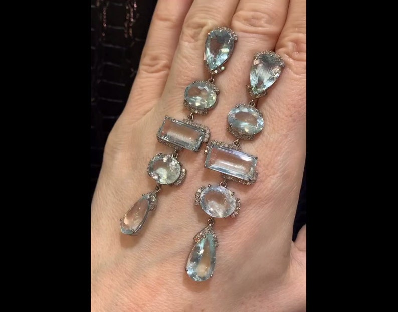 May include: A pair of long, silver dangle earrings featuring multiple aquamarine gemstones. The earrings have a variety of shapes, including pear, oval, and rectangular. The gemstones are set in silver and have a clear, light blue colour.