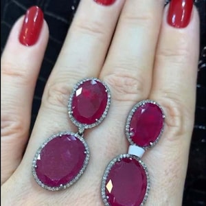 May include: A pair of dangle earrings with oval-shaped ruby gemstones surrounded by a halo of smaller diamonds. The earrings are made of silver.