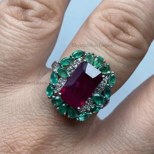 May include: A silver ring with a large, rectangular, deep red ruby, encircled by smaller, pear-shaped emeralds and tiny white gemstones. The ring is set against a light background.