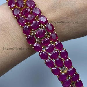 May include: A gold bracelet featuring rows of oval-cut, deep red gemstones. The bracelet has a central design with a cluster of smaller stones. The text "ShyamSilverImpex" is visible on the image.