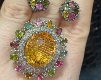 Puede incluir: Un conjunto de dos pendientes y un anillo con una gran piedra preciosa redonda amarilla rodeada por un halo de piedras preciosas blancas más pequeñas. Los pendientes y el anillo están adornados con una variedad de piedras preciosas de colores, incluyendo rosa, verde y azul.