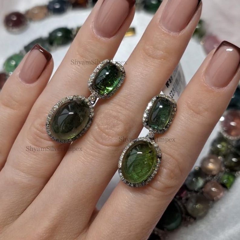 May include: A pair of dangle earrings featuring oval and rectangular green gemstones, surrounded by small, clear stones. The earrings are set in a silver metal setting. The earrings are displayed on a hand.