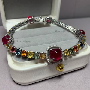May include: A silver bracelet with a series of colorful gemstones and large, square, red gemstones. The bracelet is in a white jewelry box. The text "ShyamSilverImpex" is visible on the bracelet.