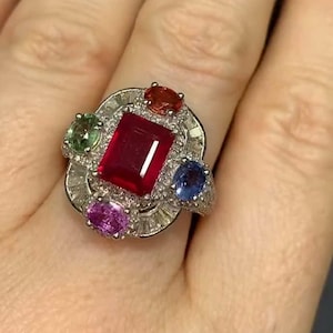 Könnte beinhalten: Ein silberner Ring mit einem großen, rechteckigen roten Edelstein in der Mitte, umgeben von kleineren, farbigen Edelsteinen in Rot, Grün, Blau und Lila. Der Ring hat einen Heiligenschein aus kleineren, klaren Steinen.