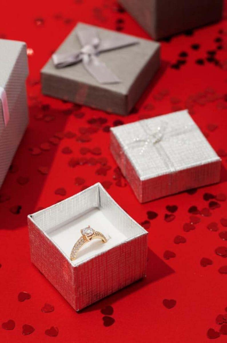 May include: A gold engagement ring with a large central stone and smaller stones on the band, displayed in an open silver gift box. Several other gift boxes with ribbons are in the background, scattered on a red surface with heart-shaped confetti.