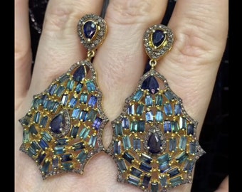 May include: A pair of gold-toned earrings with a geometric design. Each earring features a teardrop-shaped top and a larger, ornate pendant filled with blue and teal rectangular stones. The edges are lined with small, clear stones.